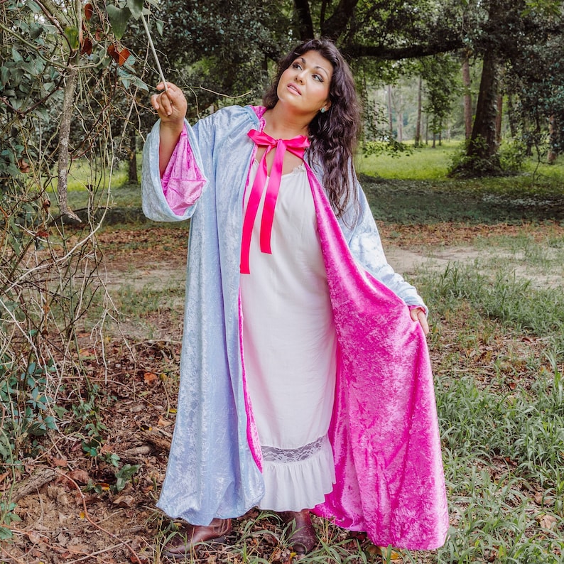 May include: A woman wearing a blue and pink velvet cape with a pink satin ribbon tied around the neck. The cape is lined with a white fabric and has a long train.