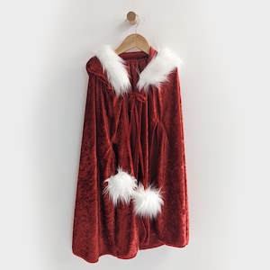 May include: Red velvet cape with white faux fur trim and a white faux fur collar. The cape has a white ribbon tie at the neck.