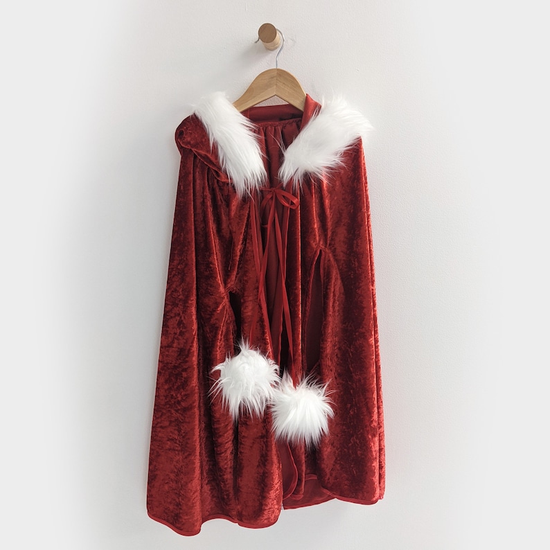 May include: Red velvet cape with white faux fur trim and a white faux fur collar. The cape has a white ribbon tie at the neck.