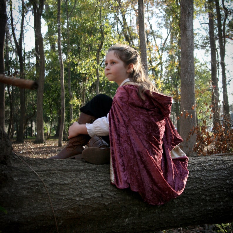 May include: A young person wearing a burgundy hooded cape sits on a fallen log in a forest. The cape has a velvet texture and is trimmed with white fabric. The person is wearing brown boots and has long brown hair.