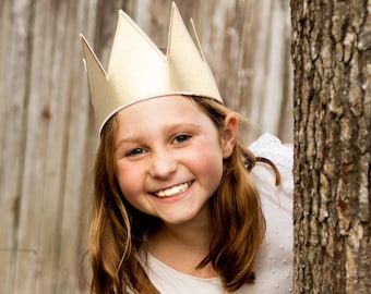 May include: A young girl wearing a gold crown smiles at the camera. She is wearing a white dress with a small pattern and has long brown hair.