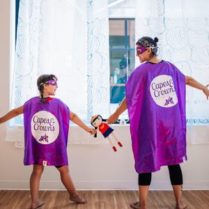 May include: Two people wearing purple capes with the logo "Capes & Crowns Foundation" are playing with a doll. The person in the back is wearing a purple bandana and the person in the front is wearing a purple mask.