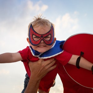 May include: A young child wearing a red superhero costume and a red and black spider-man mask is being held up in the air by an adult. The child is smiling and looking at the camera. The child is holding a red shield in their other hand.