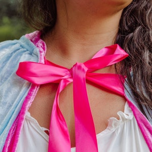 May include: A woman wearing a light blue and pink velvet cape with a bright pink satin ribbon tied around her neck in a bow.