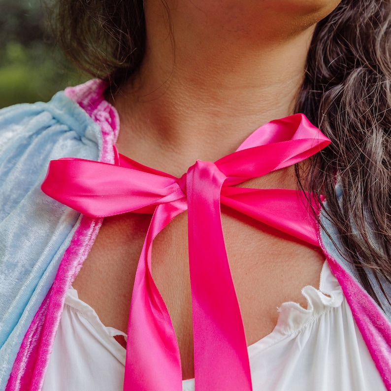 May include: A woman wearing a light blue and pink velvet cape with a bright pink satin ribbon tied around her neck in a bow.