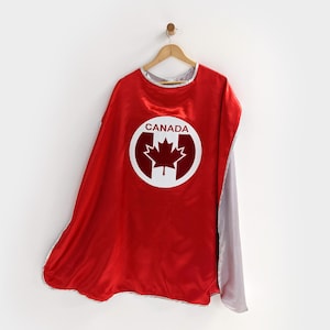 May include: A red superhero cape with a white and red Canada emblem. The cape has a silver trim and a gray lining. The cape is hanging on a wooden hanger against a white background.