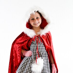 May include: A child wearing a red velvet cape with a white fur trim hood. The cape has a white fur pom-pom at the bottom. The child is wearing a black and white gingham dress and pink mittens.