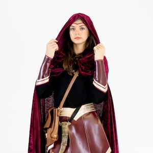 May include: A woman wearing a burgundy hooded cape with gold trim, a black long-sleeved shirt, a burgundy leather skirt, and a brown leather bag with a gold buckle. She is also wearing a gold headband with a sparkly design.