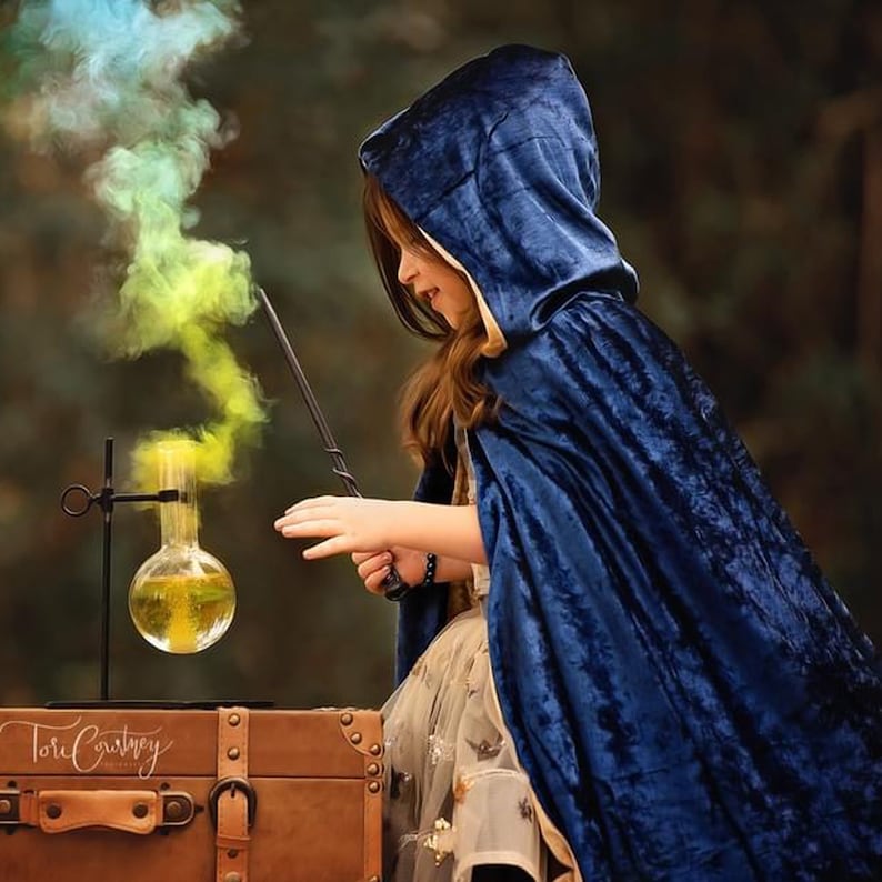 May include: A young person wearing a blue velvet hooded cape holds a wand and points it at a glass flask with yellow liquid, creating a colorful smoke effect.