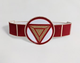May include: Red and white headband with a gold glitter embroidered triangle design. The headband is made of felt and has a white background with a red triangle in the center. The triangle has gold glitter embroidery.