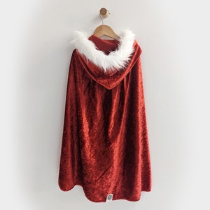 May include: Red velvet hooded cape with a white faux fur trim. The cape is hanging on a wooden hanger against a white wall.