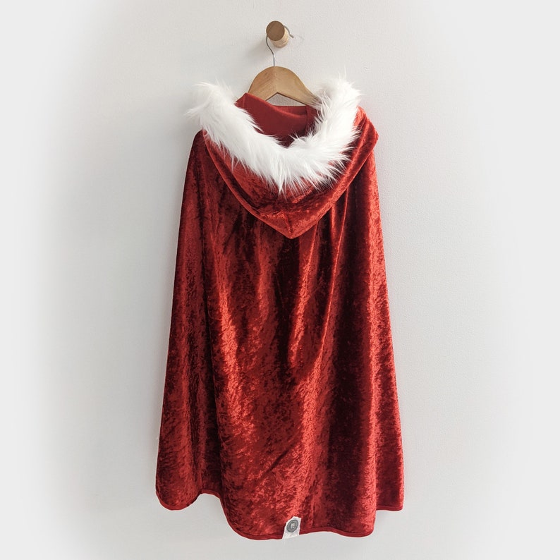 May include: Red velvet hooded cape with a white faux fur trim. The cape is hanging on a wooden hanger against a white wall.