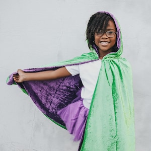 May include: A young person wearing a green and purple hooded cape with a white shirt and a purple tulle skirt. The cape has a velvet texture and is lined with a purple fabric.