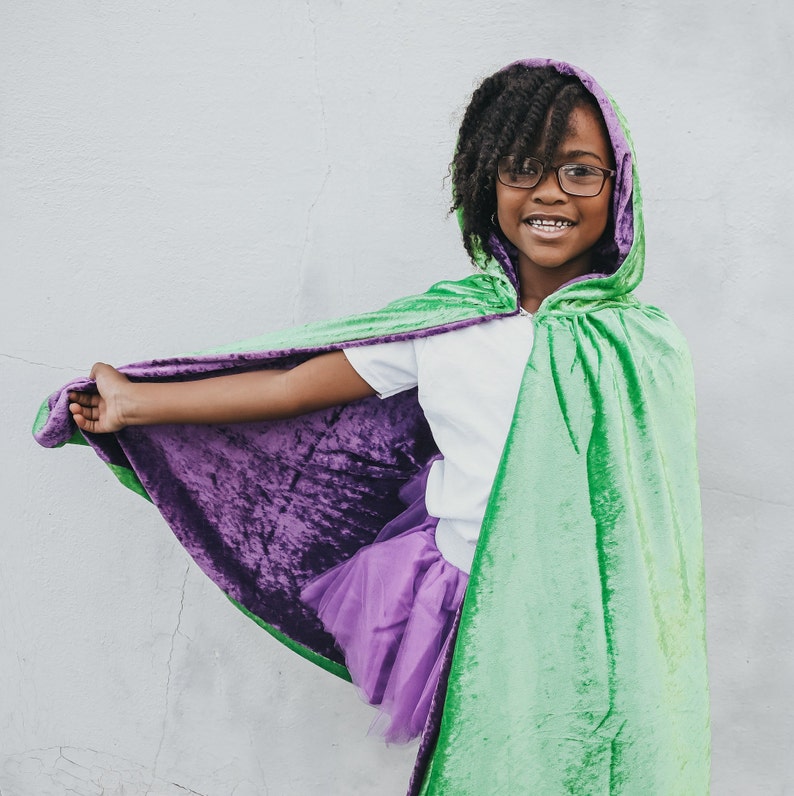 May include: A young person wearing a green and purple hooded cape with a white shirt and a purple tulle skirt. The cape has a velvet texture and is lined with a purple fabric.