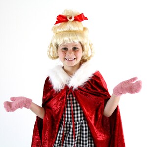 May include: A young person wearing a red velvet cape with a white fur collar, a black and white checked dress, and pink mittens. The person is wearing a blonde wig with a red bow on top.