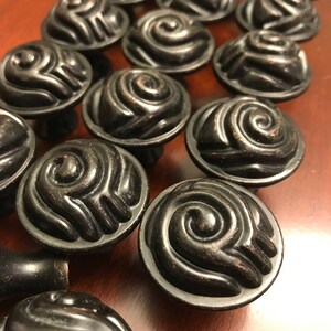May include: A collection of round cabinet knobs featuring a dark bronze finish and a raised, swirling spiral pattern on the face. Each knob has a circular base and a smooth, curved top surface with deep grooves that create a textured, dimensional look.