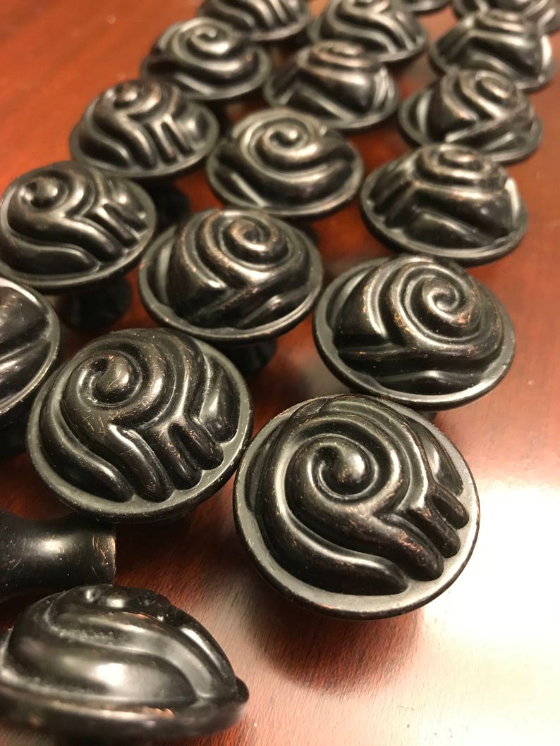 May include: A collection of round cabinet knobs featuring a dark bronze finish and a raised, swirling spiral pattern on the face. Each knob has a circular base and a smooth, curved top surface with deep grooves that create a textured, dimensional look.