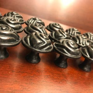 May include: A collection of black cabinet knobs featuring a raised, swirling rose-like pattern on the circular top. Each knob has a smooth, rounded base and a dark, matte finish with subtle metallic highlights visible on the curved edges.