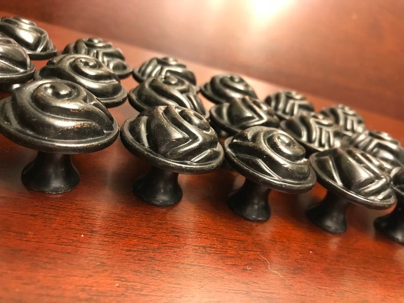 May include: A collection of black cabinet knobs featuring a raised, swirling rose-like pattern on the circular top. Each knob has a smooth, rounded base and a dark, matte finish with subtle metallic highlights visible on the curved edges.