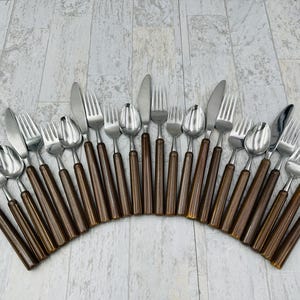 May include: A collection of stainless steel silverware, including forks, knives, and spoons, arranged in an arc. The silverware features brown handles. The arrangement is on a light gray, wood-grain surface.