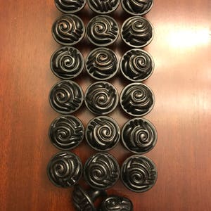 May include: A collection of eighteen round cabinet knobs arranged in a grid, plus two additional knobs placed at the bottom. Each knob is dark black with a metallic bronze-toned finish on the raised spiral swirl pattern on the circular face.