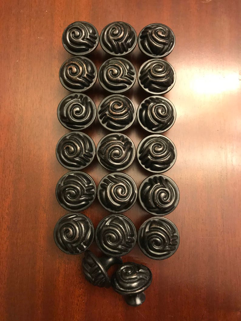 May include: A collection of eighteen round cabinet knobs arranged in a grid, plus two additional knobs placed at the bottom. Each knob is dark black with a metallic bronze-toned finish on the raised spiral swirl pattern on the circular face.