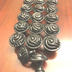 May include: A collection of dark bronze-toned metal cabinet knobs arranged in a grid. Each knob features a circular top with a raised, swirling spiral pattern that resembles a stylized rose or shell. The knobs have a smooth, rounded base.