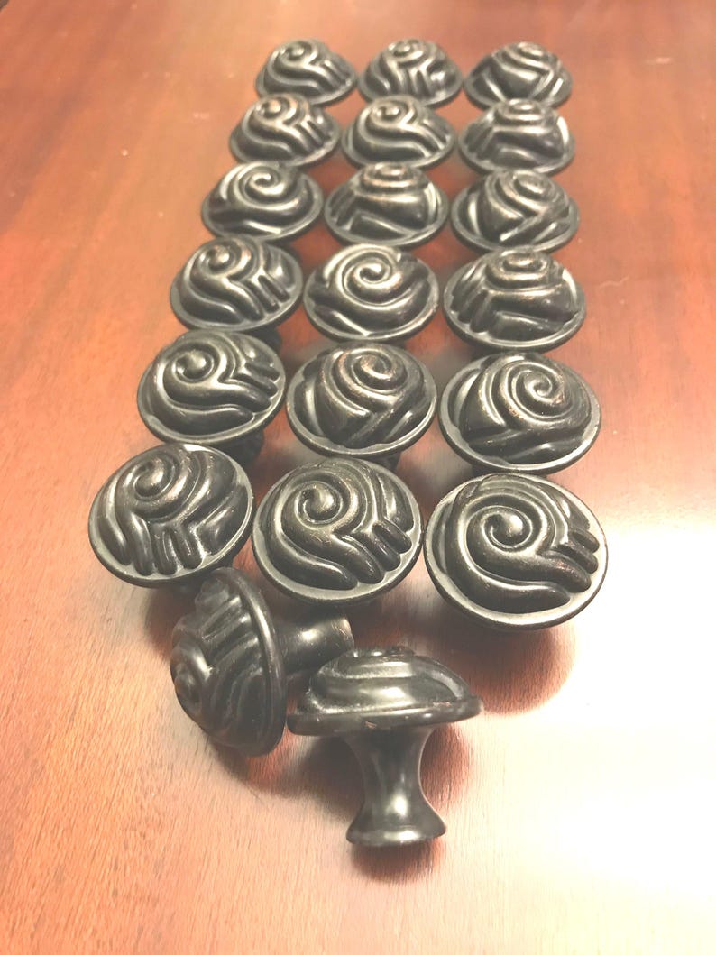 May include: A collection of dark bronze-toned metal cabinet knobs arranged in a grid. Each knob features a circular top with a raised, swirling spiral pattern that resembles a stylized rose or shell. The knobs have a smooth, rounded base.