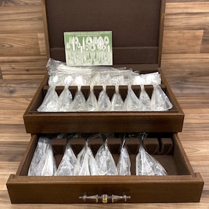 May include: A wooden silverware chest with two drawers, each containing a set of silverware wrapped in plastic. The chest has a brass handle and a label that reads "Complete Your Service With Oneida Open Stock".