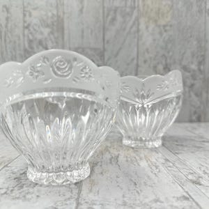 May include: Two clear glass bowls with a scalloped rim. The rim of the larger bowl has a frosted floral design. The bowls have a textured, ribbed design on the sides. The bowls are sitting on a light gray wooden surface.
