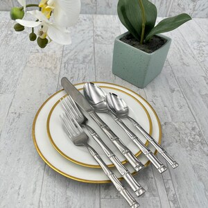 May include: A set of five silver bamboo-style flatware pieces, including a knife, fork, and three spoons, on a white plate with a gold rim.