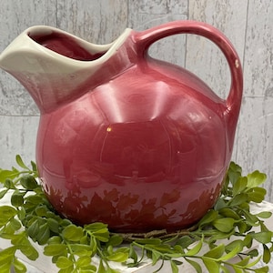 May include: A vintage red ceramic pitcher with a white rim. The pitcher has a round body and a curved handle.
