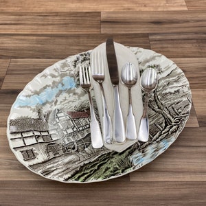 May include: A white oval platter with a brown and green floral design featuring a cottage and a horse-drawn cart. The platter has a scalloped edge and is decorated with a blue and white sky. A set of silver cutlery is placed on top of the platter, including a knife, fork, and two spoons.