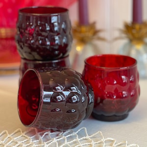 May include: A collection of four ruby red drinking glasses. The glasses have a textured, dimpled surface and a rounded shape. One glass is lying on its side, while the others are stacked or upright. The glasses are on a white surface.