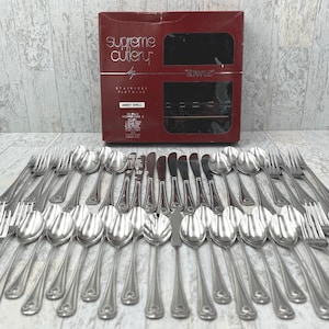 May include: A complete set of stainless steel flatware, including forks, spoons, and knives, arranged in rows. The set is from "Supreme Cutlery" and includes a red box with the "Abbey Shell" pattern.