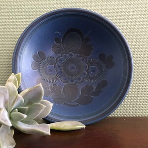 May include: A round, deep blue ceramic bowl featuring a central embossed floral pattern with intricate petal and leaf details. The bowl has a smooth, matte finish and a slightly raised rim, showcasing a dark blue hue throughout the interior surface.