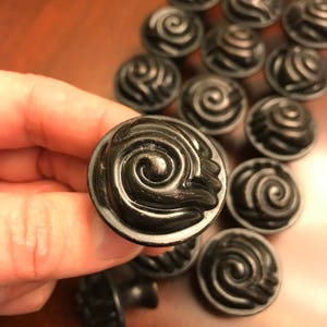 May include: A collection of round, dark bronze-colored cabinet knobs featuring a raised, swirling spiral pattern on the face. Each knob has a circular base and a smooth, curved edge, with a metallic finish that shows subtle highlights on the ridges.