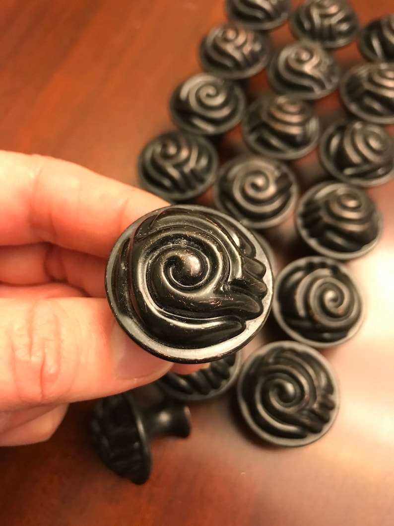 May include: A collection of round, dark bronze-colored cabinet knobs featuring a raised, swirling spiral pattern on the face. Each knob has a circular base and a smooth, curved edge, with a metallic finish that shows subtle highlights on the ridges.
