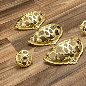 May include: Four gold-toned metal cabinet knobs with a decorative, intricate design. Two of the knobs are larger and have a curved shape, while the other two are smaller and round.