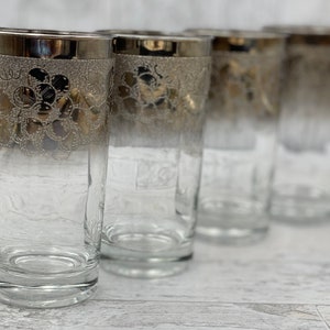 May include: Set of four clear glass tumblers with a silver rim and a floral design. The glasses have a frosted effect at the top and are clear at the bottom.