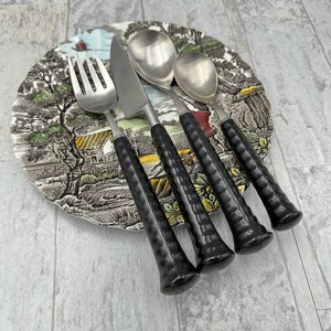 May include: A set of four silver-toned flatware pieces with black, textured handles. The set includes a fork, knife, and two spoons.