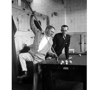 Peut inclure: Photographie en noir et blanc de deux hommes jouant au billard. L'un est assis sur le bord de la table, visant avec une queue. L'autre homme regarde. Plusieurs boules de billard sont sur la table en feutre vert.