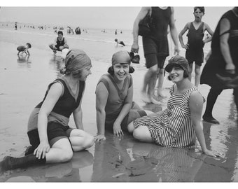 May include: Three women in vintage swimwear pose on a beach. Two women are wearing matching red and black swimsuits with matching headbands. The third woman is wearing a striped swimsuit and a black and white hat.