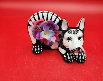 May include: A black and white skeletal dog figurine with a bouquet of pink, purple, and blue flowers. The dog is lying down with its head resting on its paws.