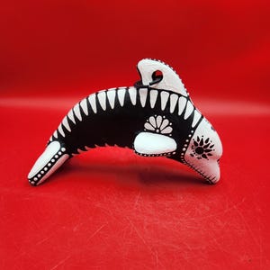 May include: A hand-painted, decorative orca figurine. The orca is black and white with floral and dot patterns. The figurine has a loop at the top for hanging. The background is a solid red.