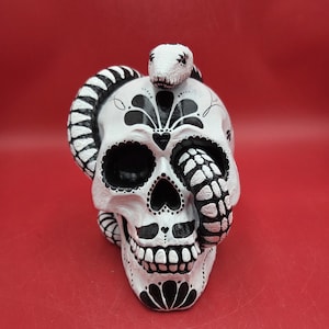 May include: A white skull figurine with black decorative patterns and a snake wrapped around it. The snake's head rests on top of the skull. The skull has large eye sockets and a detailed jawline. The background is red.