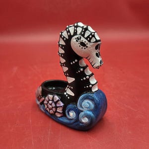 May include: A Day of the Dead seahorse figurine. The seahorse is black with white accents and a skull-like face. The base is blue with white swirls and a floral design. The figurine is a unique decorative item.