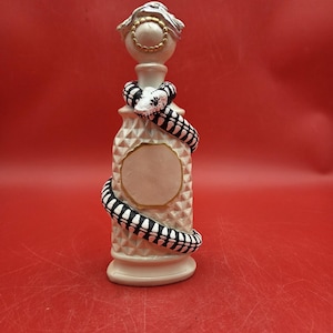 May include: A decorative perfume bottle with a white and gold stopper. A black and white snake is wrapped around the bottle. The bottle has a diamond pattern and a blank oval space.