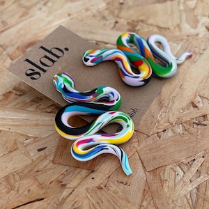 May include: Three colourful, abstract snake sculptures with a marbled effect. The snakes are made of various colours, including green, blue, yellow, and pink. The word "slab." is printed on a brown tag in the background.