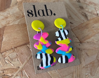 Puede incluir: Un par de pendientes abstractos y coloridos con una parte superior amarilla, formas rosas, azules y amarillas y elementos a rayas blancas y negras. Los pendientes están sujetos a una tarjeta marrón con la palabra "slab."